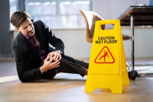 A man lying on the ground is clutching his knee after a fall. A yellow wet floor sign is in the foreground.