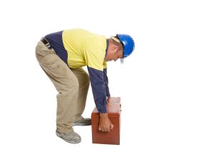 A man wearing a blue hat bending down to pick up a wooden box using the incorrect lifting technique. 