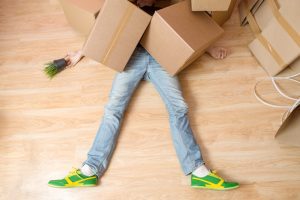 Man with blue jeans and green shoes lying on wooden floor with boxes covering his upper body. 