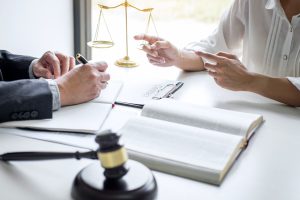 A desk, upon which are two notepads, a gavel, justice scales, and the hands of both a workplace accident solicitor and their client. They are discussing claiming for injuries due to no training at work.