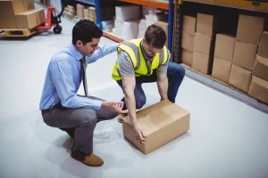 A manager in a shirt and tie kneels next to a worker who is picking up a box.
