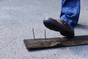 Two exposed nails sticking out of a wooden board about to be stepped on by a work boot