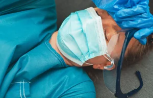 A medical professional lying on the floor wearing a face mask and blue scrubs. 