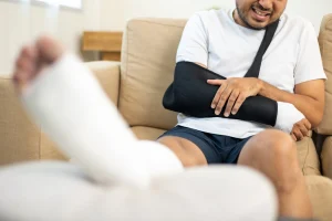 A man wearing a sling holding his arm in pain while his leg in a cast is propped up on the sofa.