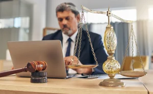 A solicitor writing about public liability claims on his laptop sat at a desk that has a wooden gavel and gold and glass scales.